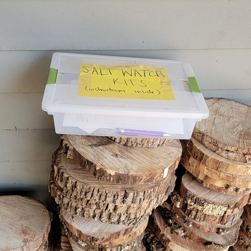 Plastic container with a label that reads "Salt Watch Kits (instructions inside). The container is sitting on top of a stack of wood cookies leaning against the side of a building.