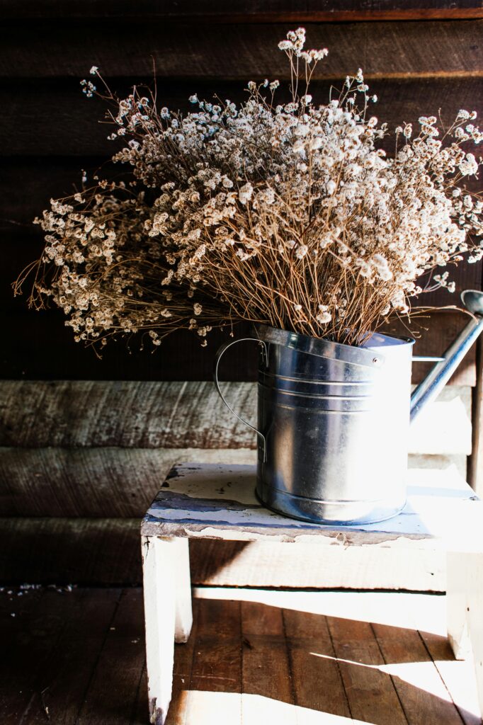 Dried flowers arranged in a metallic watering can on a white wooden stool in natural light. I much prefer using metal watering cans over plastic which tend to crack and puncture with repeated use. A recyclable metal can that can last a lifetime is a simple way to consider long-term sustainability when it comes to every-day tools.